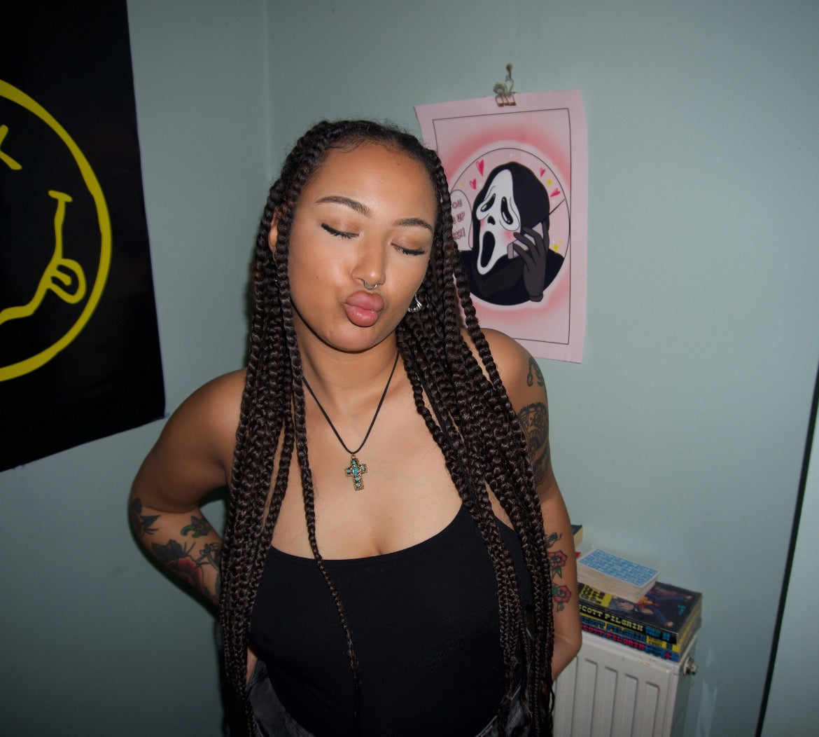 a girl standing in a bedroom pouting, she's wearing a gold and turquoise vintage-inspired cross pendant on a black cord chain