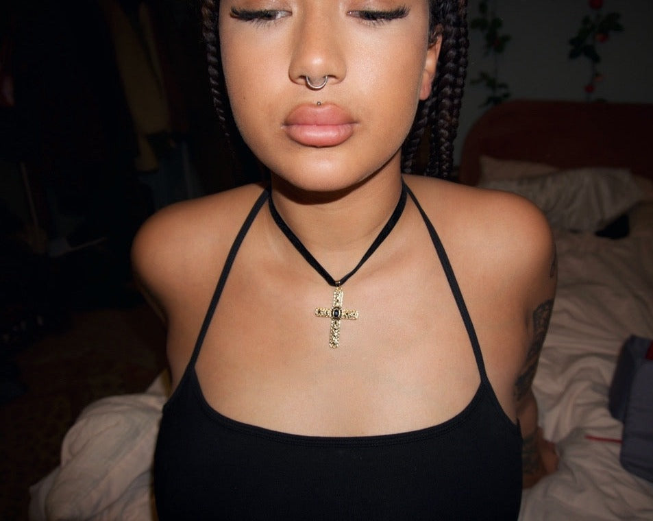 an up close shot of a girl sat on the end of a bed wearing a black velvet choker with a gold cross pendant adorned with black gems