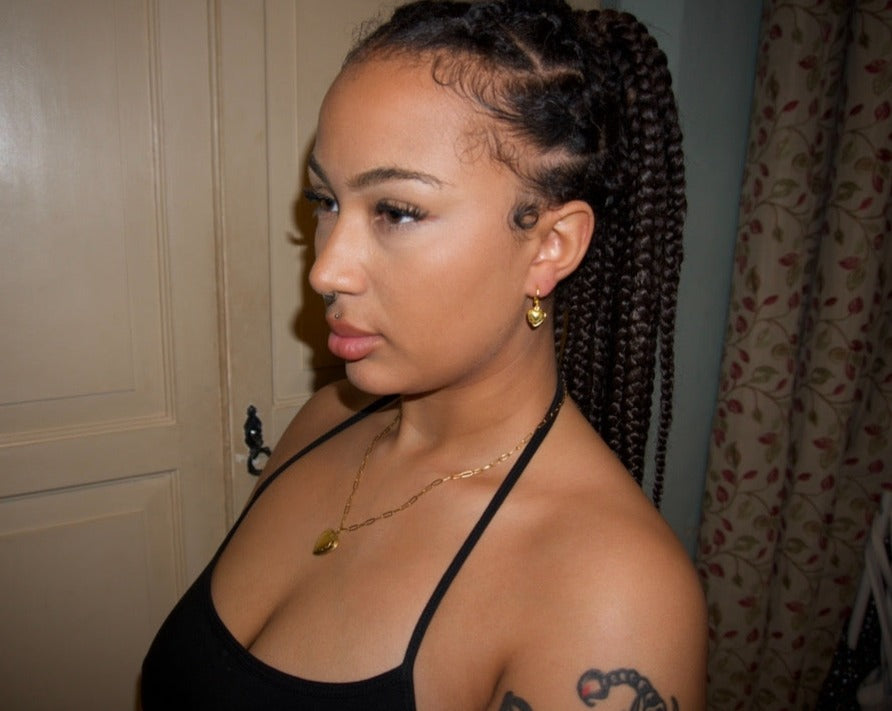 a girl with a braided ponytail looking to the side showing off her jewellery. she wears a dainty gold chain with a detailed heart shaped locket pendant, with a matching pair of gold locket heart earrings