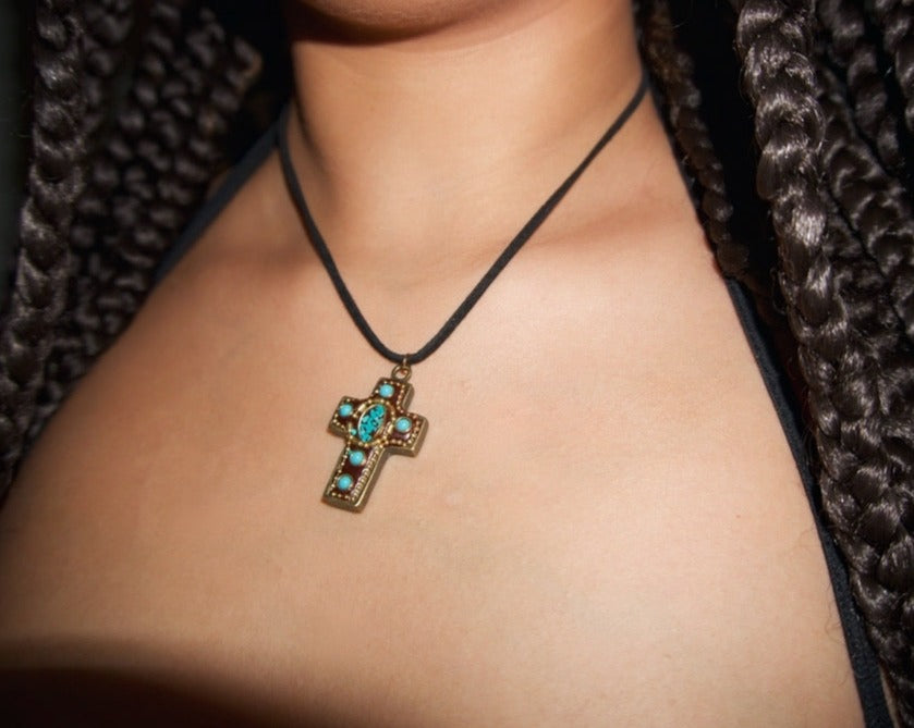 a close up shot of a girl wearing a gold and turquoise vintage-inspired cross pendant on a black cord chain