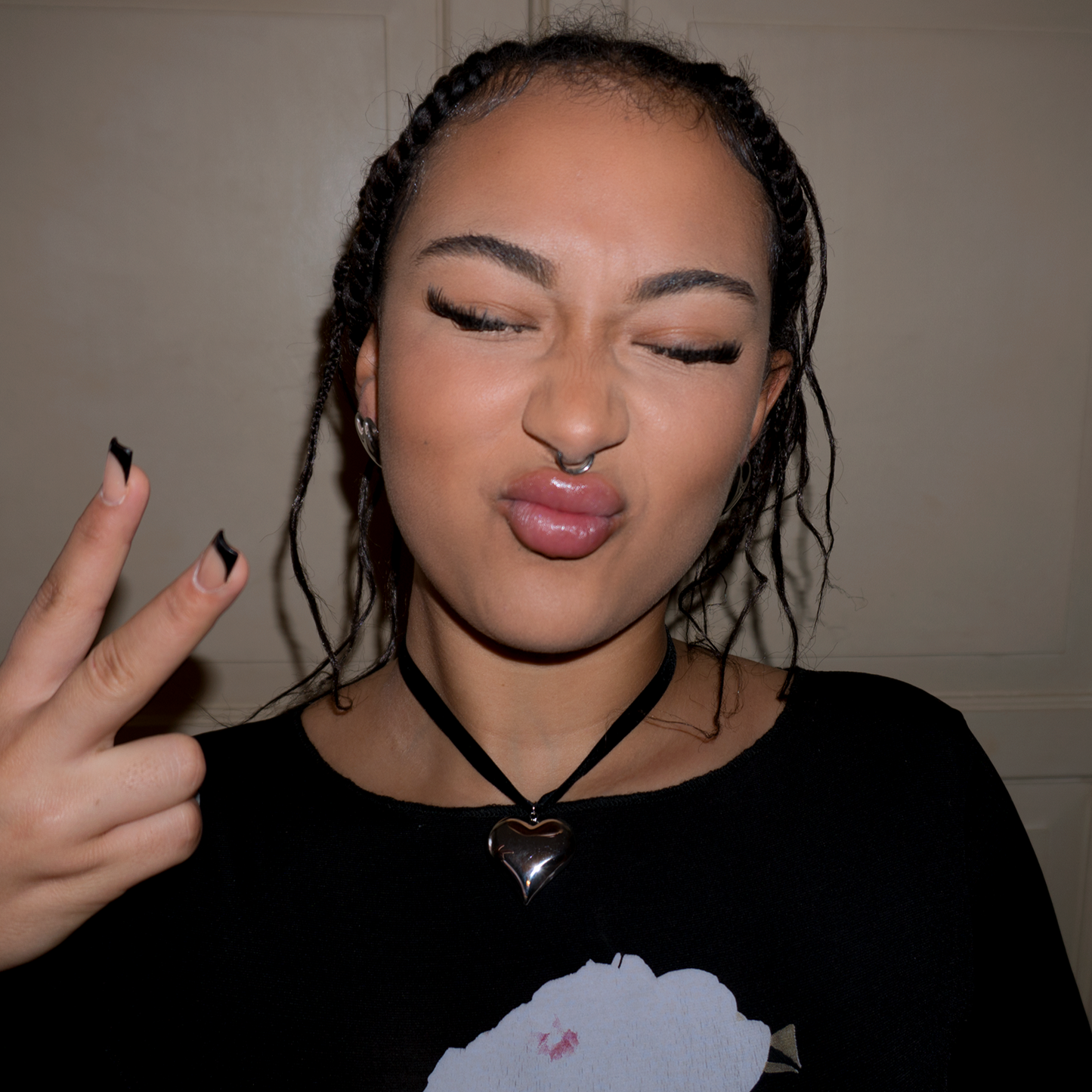 A girl with black and pink nails doing the peace sign and pouting. She’s wearing a black ribbon choker with a silver heart pendant on it.