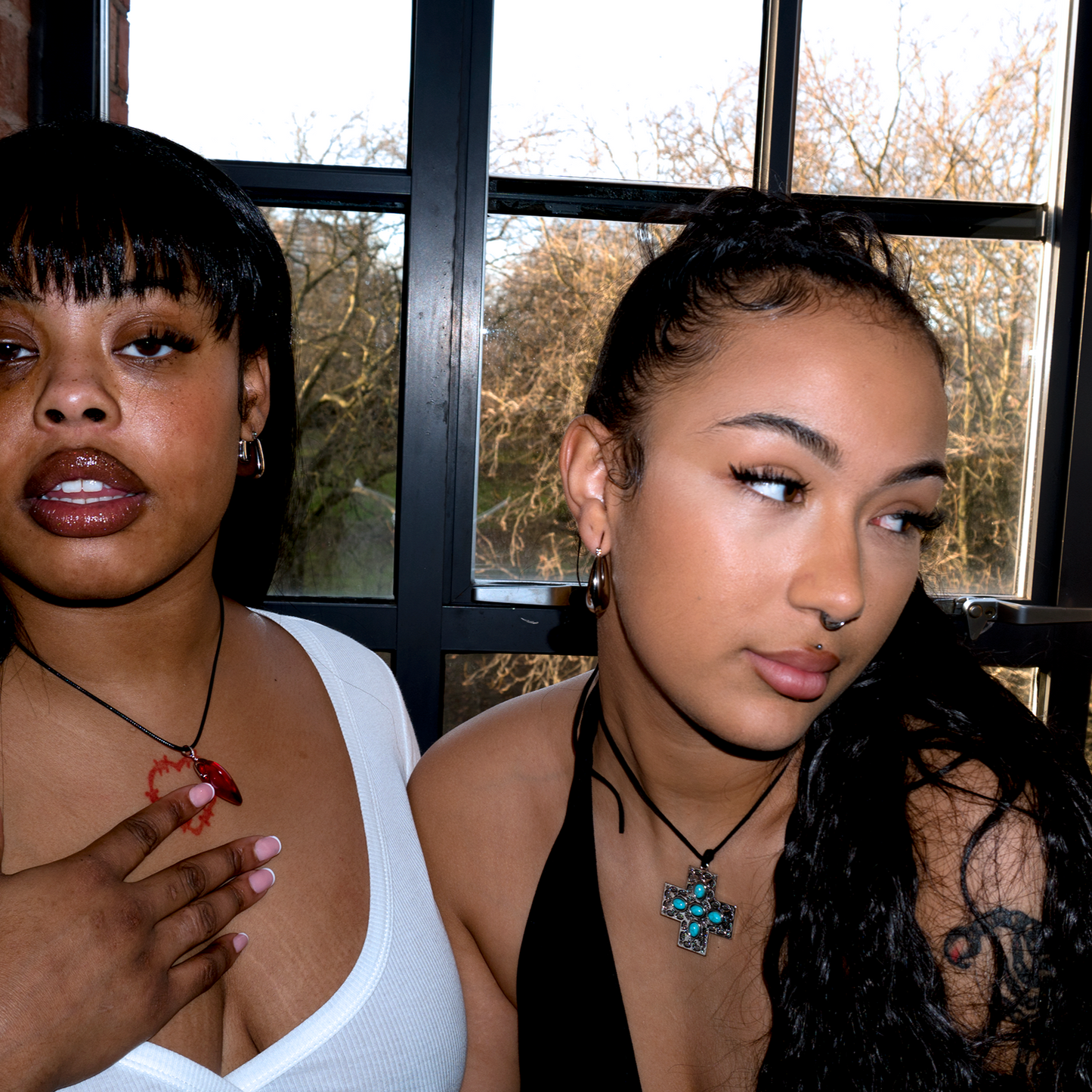 2 models sat in front of a window, one is wearing a chunky red heart necklace and the other wears a silver cross necklace with blue beads in the centre of the cross