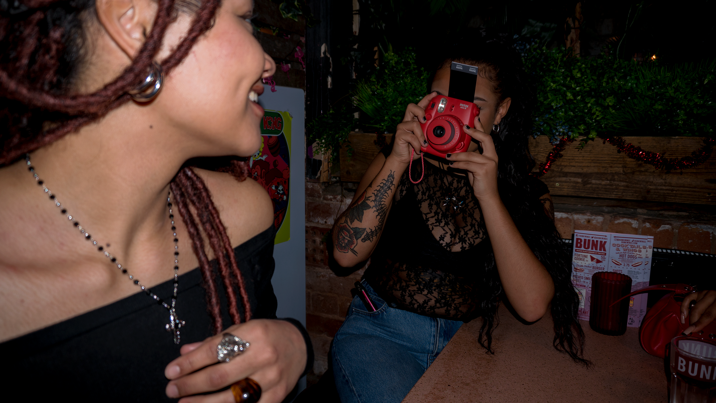 A woman taking a picture on a Polaroid camera of another woman wearing a silver rosary cross necklace