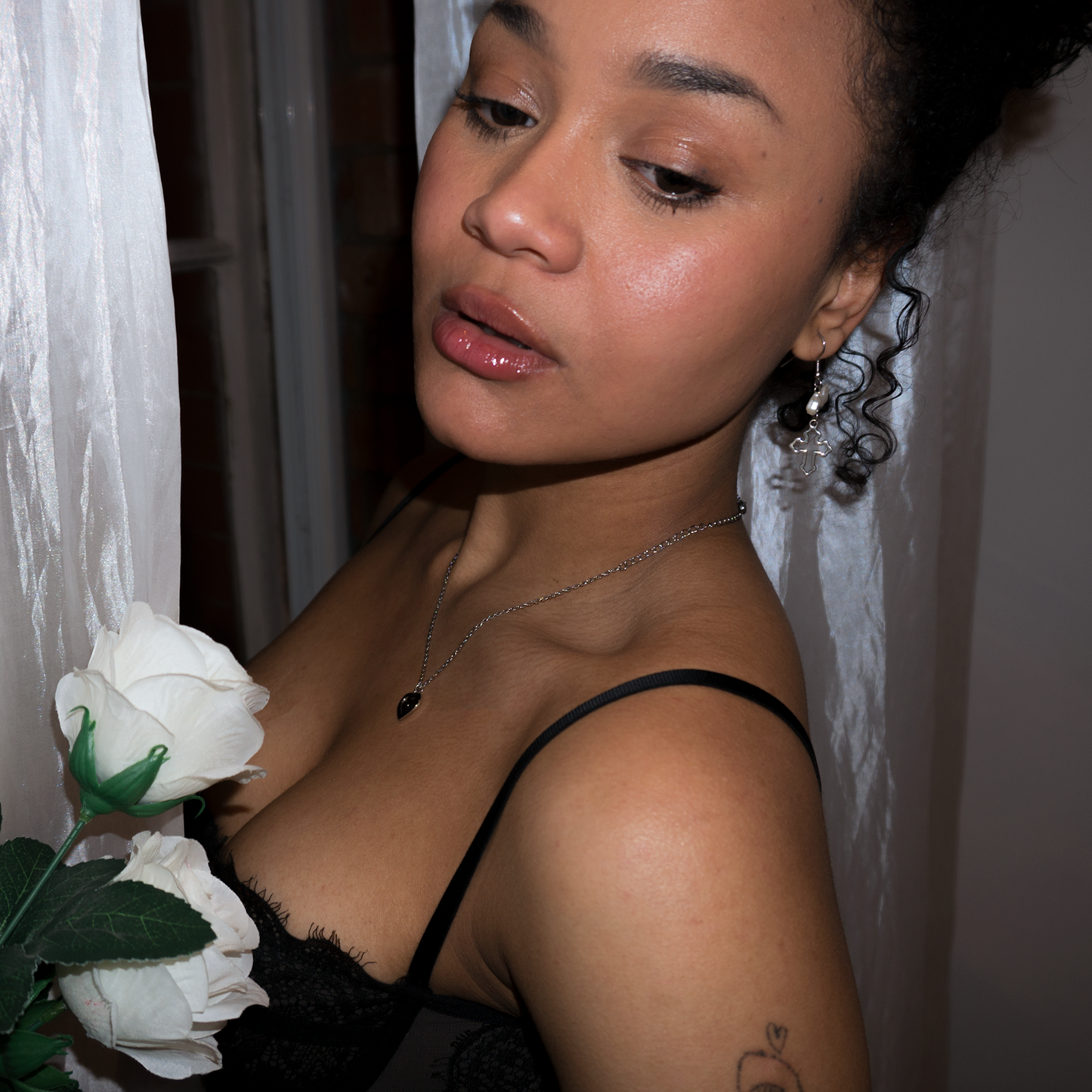 a girl holding white roses standing in front of a white satin curtain wearing dangly cross earrings anda dainty rhinestone star engraved heart pendant