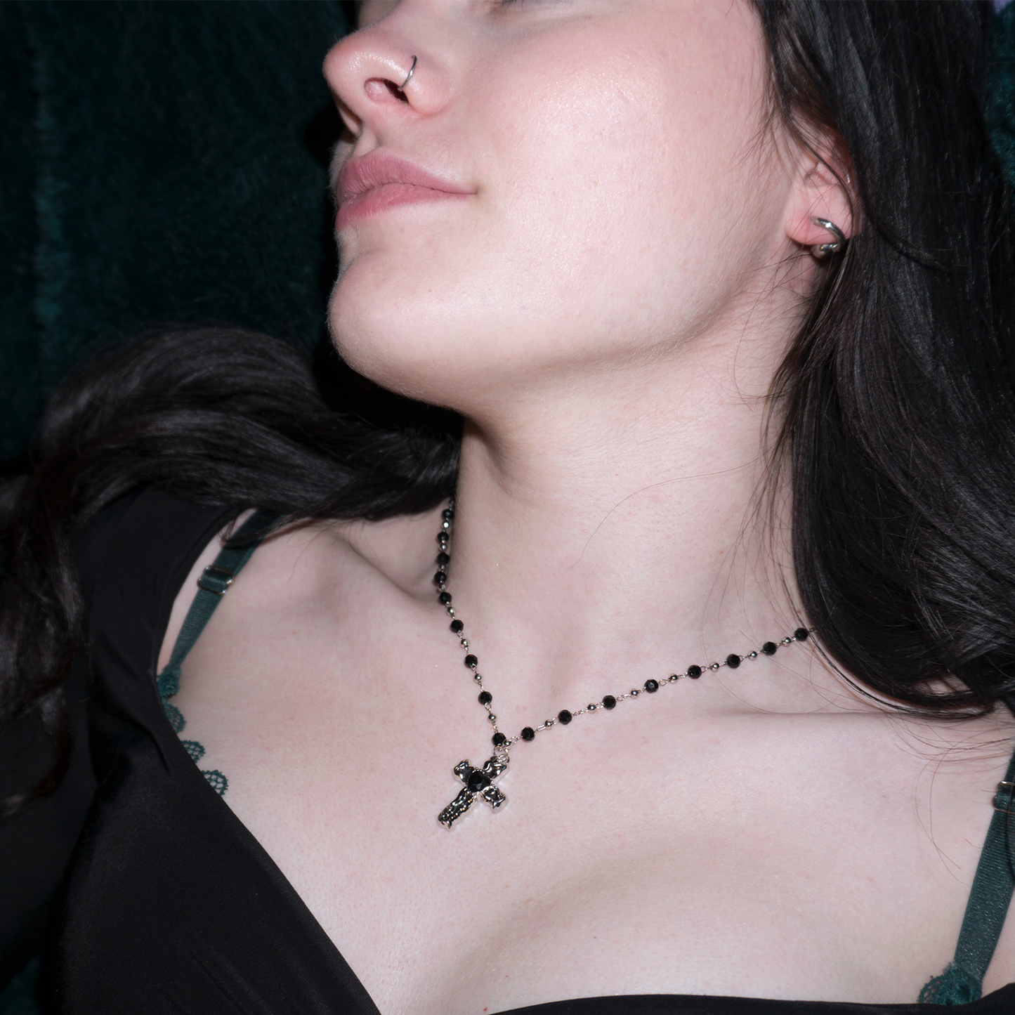 girl with black long hair laying back on a dark green fur blanket wearing a black and silver rosary inspired necklace, featuring a cross pendant