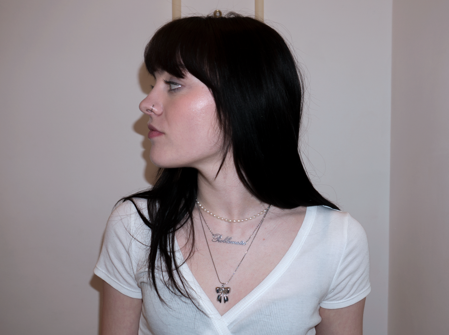 a girl with black hair and a fringe wearing layered necklaces with pearls. one necklace is a bow pendant and the second one is a 'Problematic' nameplate necklace