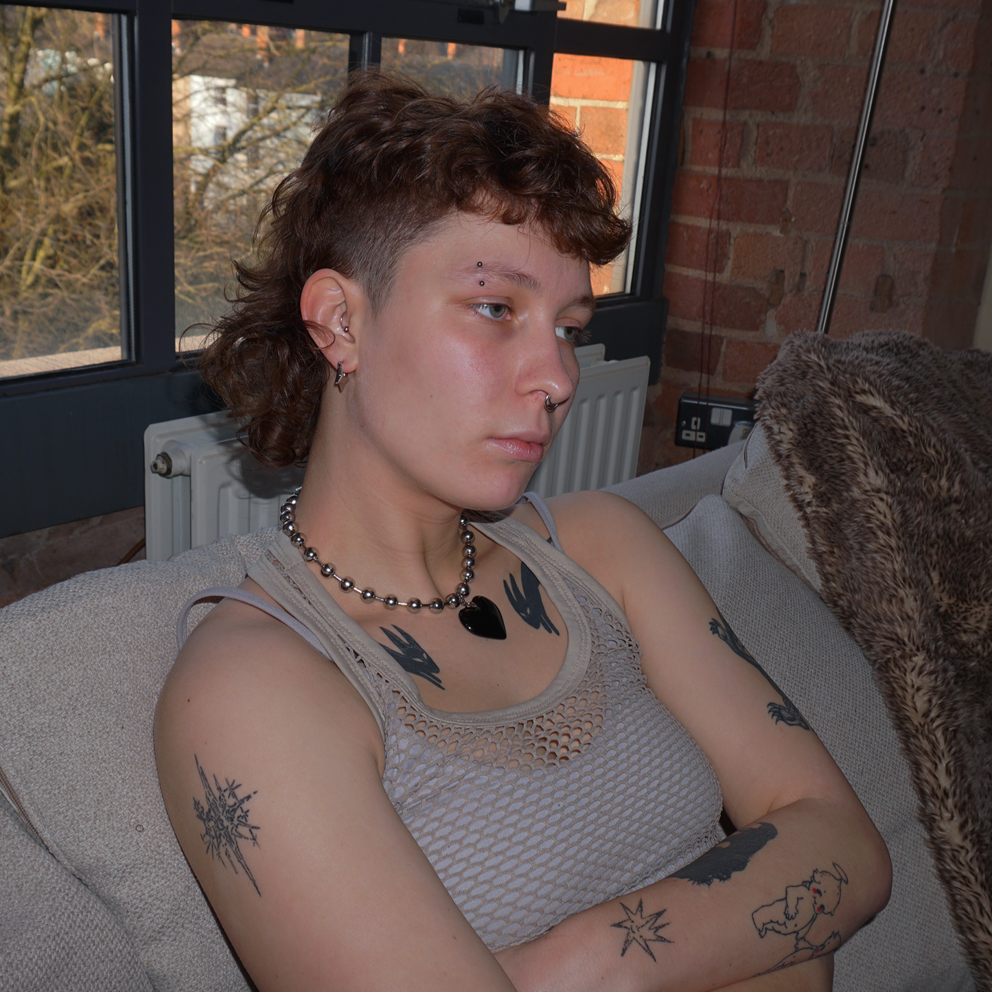 Model sat casually on a sofa wearing a chunky metal ball chained necklace, with a large black heart pendant on
