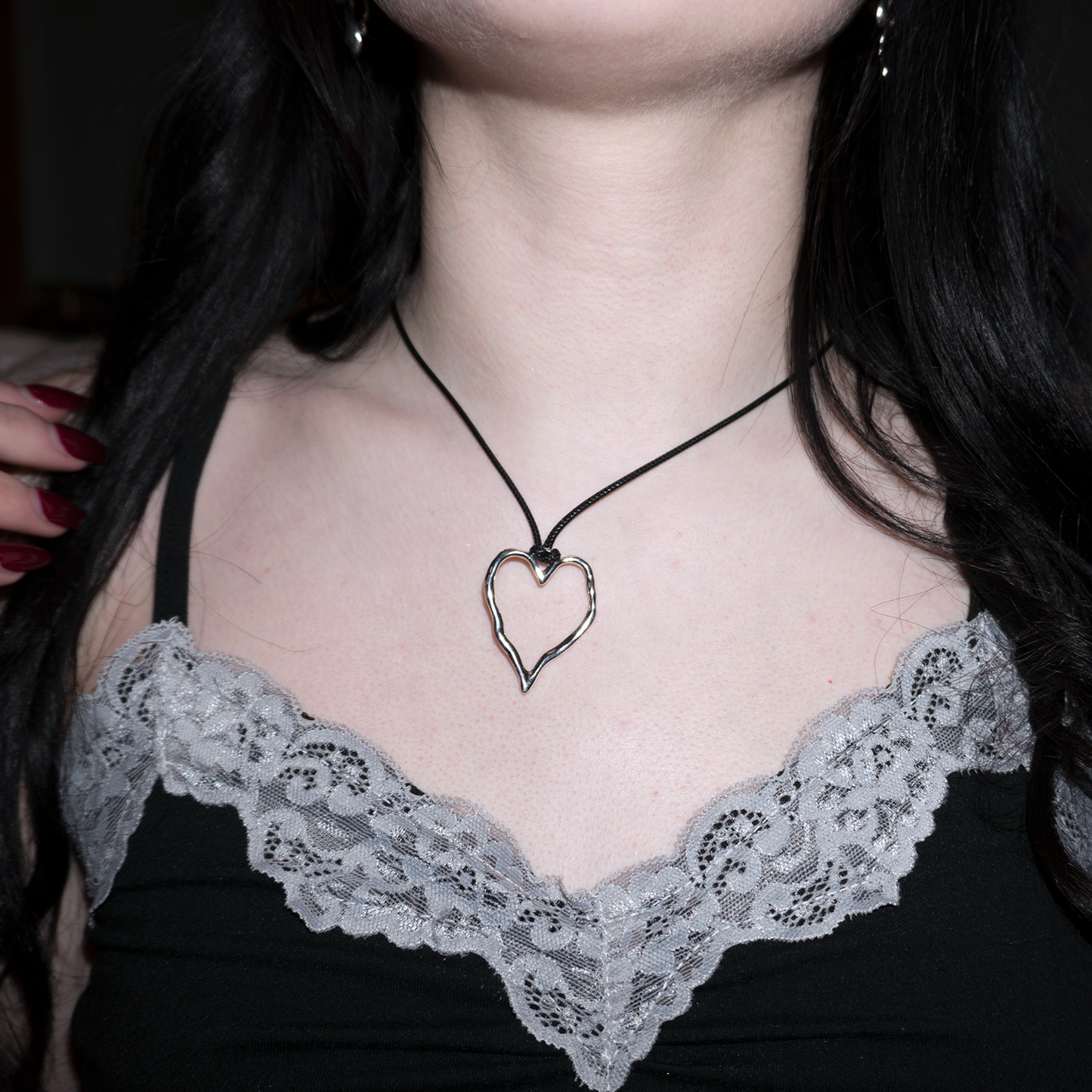 girl with red nails brushing her hair back to reveal her necklace. its a black leather cord chain with a stainless steel silver heart pendant in a hammered shape
