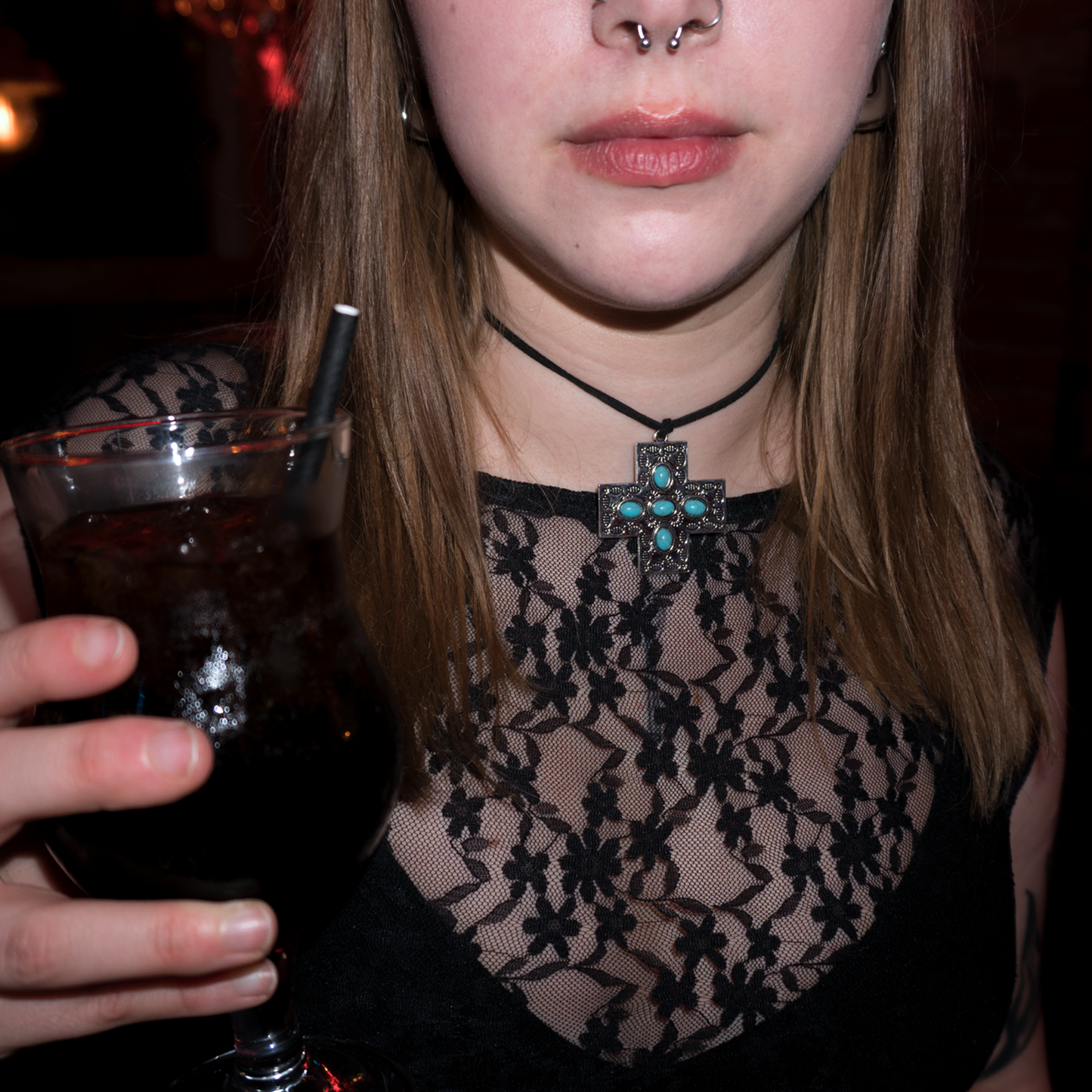Close up of models neck as she holds a glass with a dark drink in it. She is wearing a silver cross pendant with 5 blue beads inside it