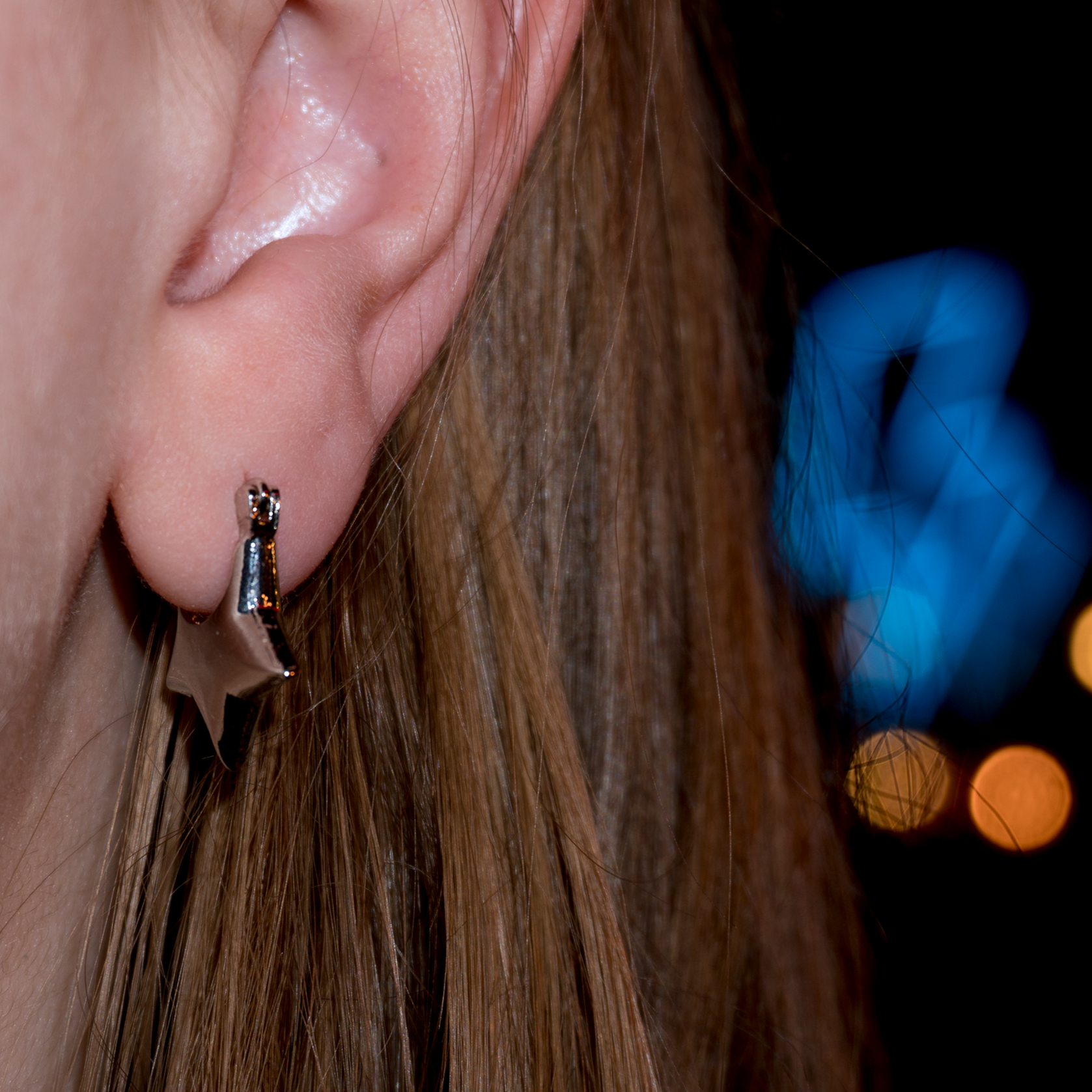 Extreme close up of a models ear wearing a small start shaped pendant, there are blurred lights in the background