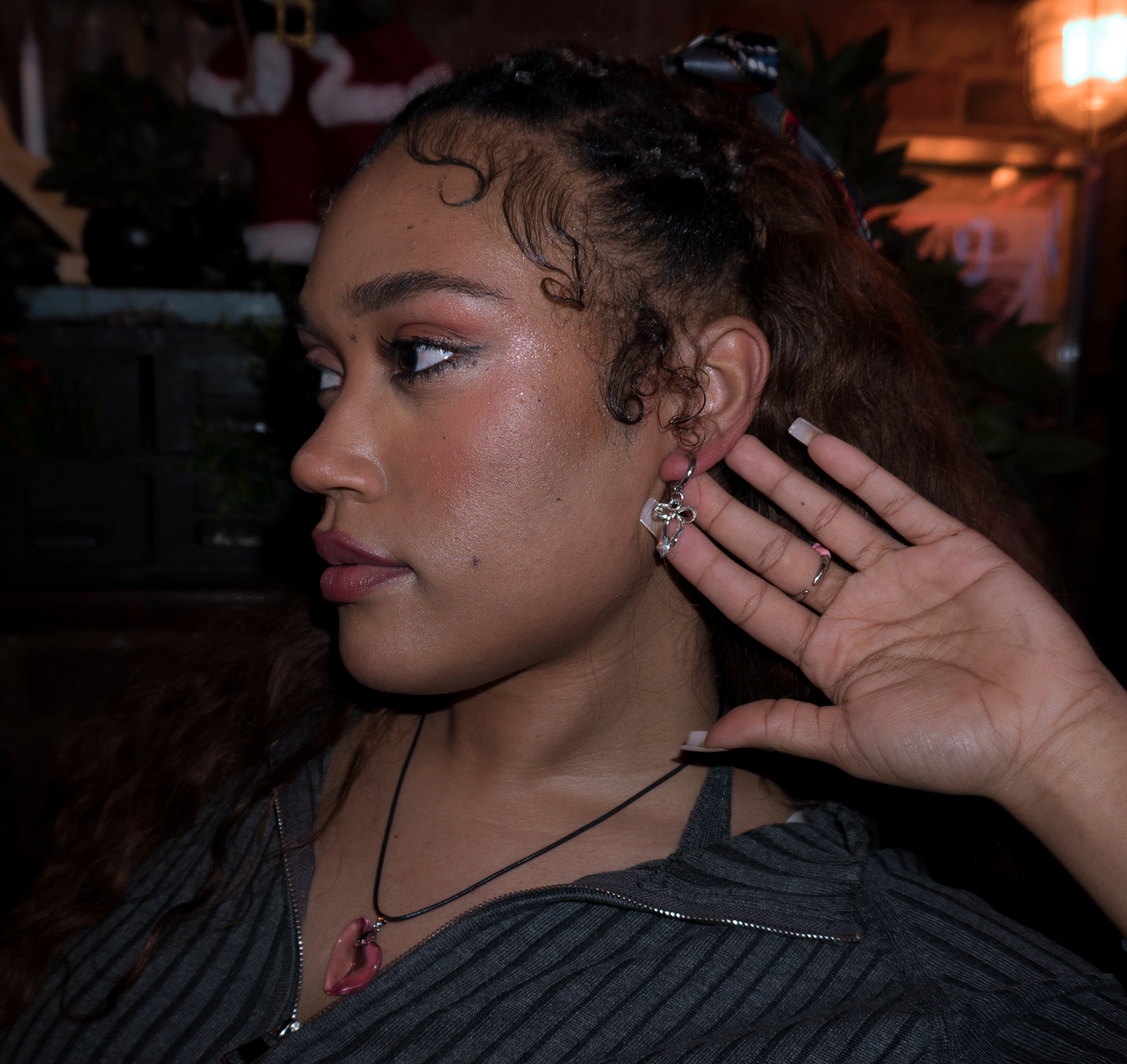 A black model with acrylic nails and styled baby hair, she’s got light brown curly hair and a heart shaped freckle on her cheek. The girl is wearing a pink glass heart necklace on a black cord chain and showing off her dangly bow shaped pendant hoop earrings