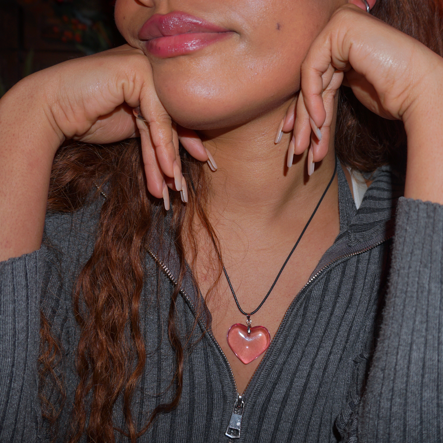 Close up of model wearing a grey jacket, it is slightly up zipped to reveal a shiny pink glass heart pendant