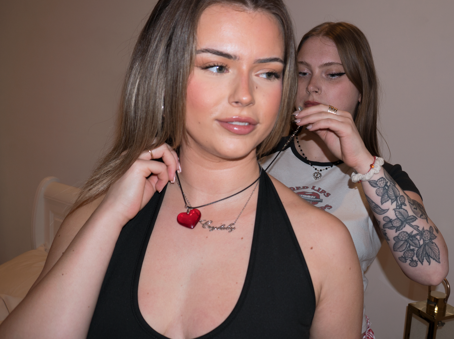 two blonde girls, one helping the other one put on a glass red heart necklace. the girl in front is smiling and wearing a 'Crybaby' nameplate necklace