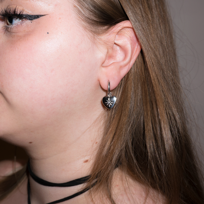 a girl with mousy brown hair looking to the side, the camera is focusing on her hoop earrings with a dangly heart pendant on them, they have stars engraved on the front